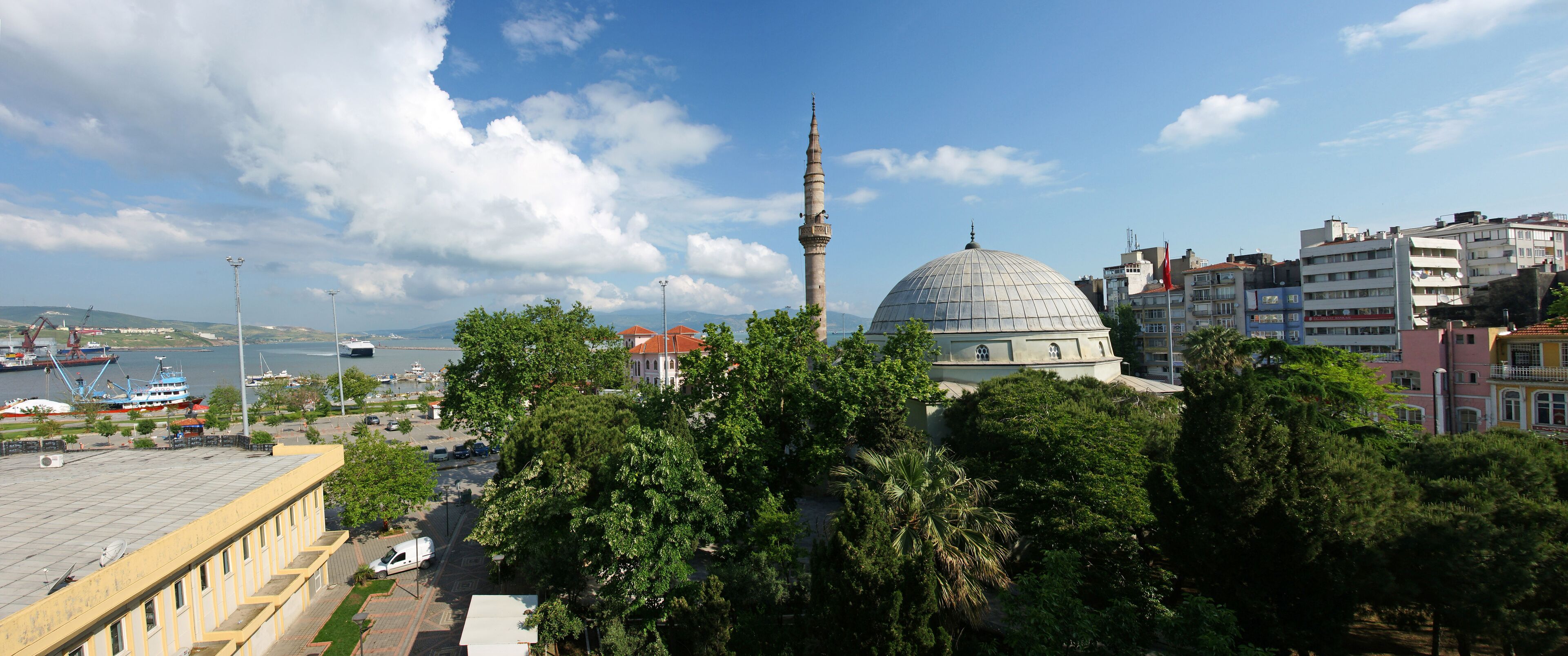 A cityscape with a prominent mosque featuring a tall minaret and a large dome, surrounded by lush green trees and modern buildings.