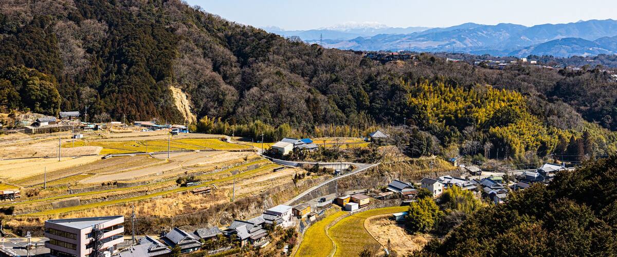 A rural landscape with a winding road, buildings, and a forested hill with distant mountains.