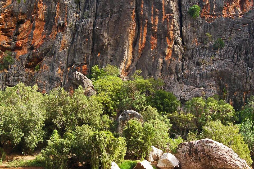 A river reflecting tall cliffs with red rock formations and green trees.
