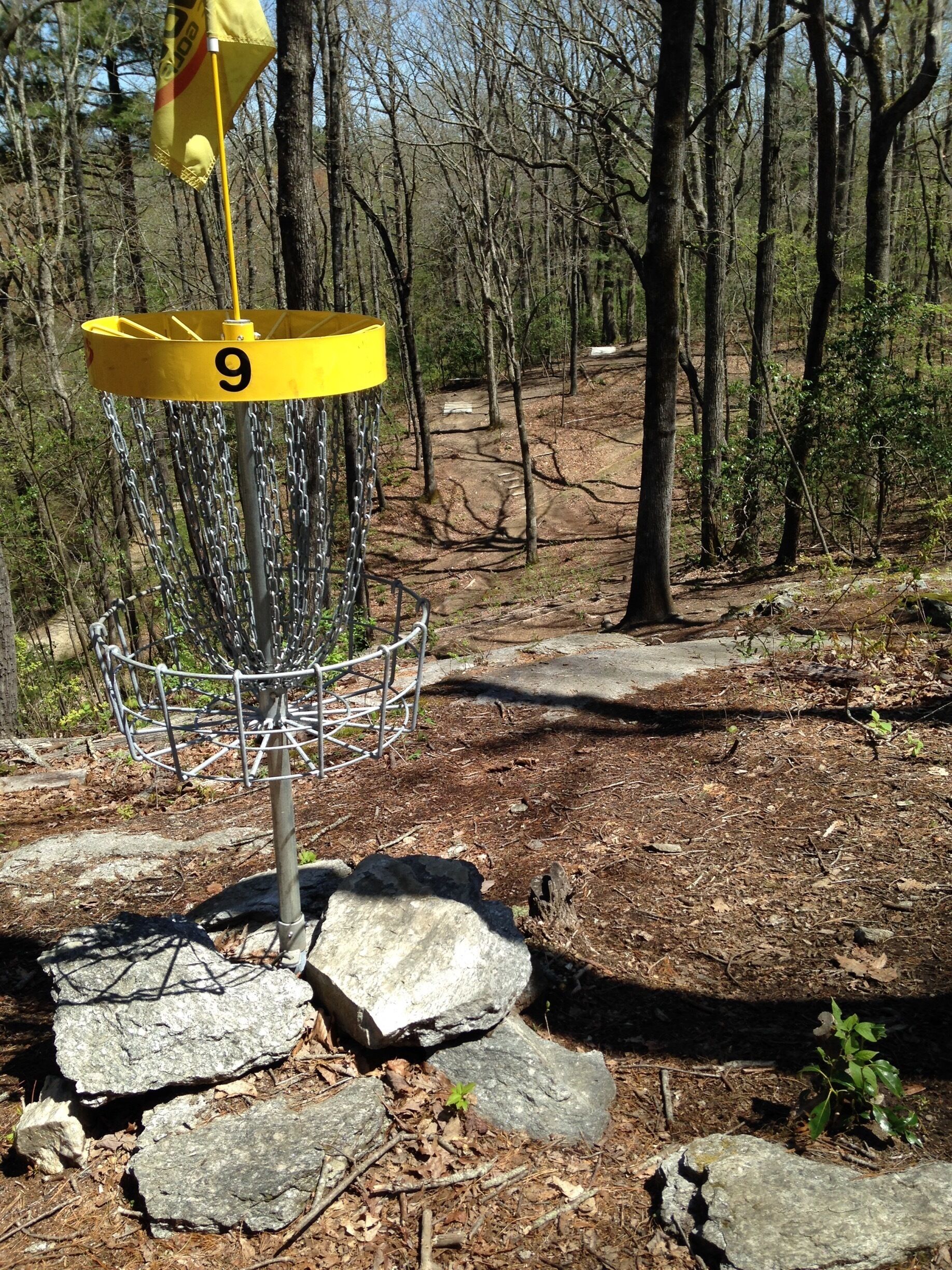 A disc golf basket labeled '9' in a wooded area with a dirt path and scattered rocks.