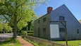 A historical homestead with a wooden exterior, a brick chimney, and a signboard detailing its history.
