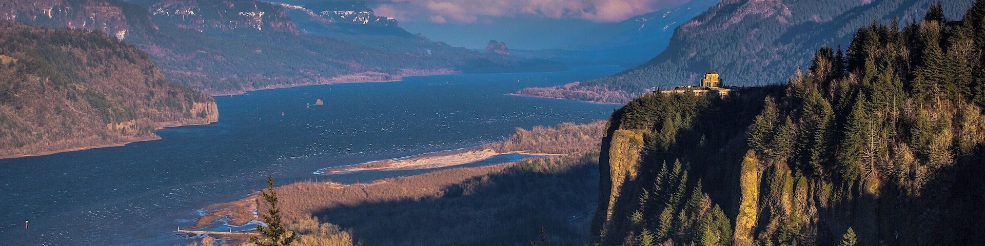 A mountainous landscape with a lake, forested areas, and a building on a cliff.