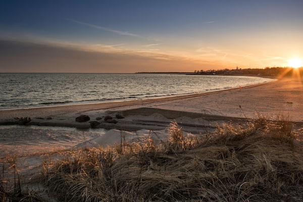 Ein Strand bei Sonnenuntergang, die Sonne tief am Horizont, ruhiges Wasser und grüne Dünen.