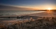 Une plage au coucher du soleil, avec le soleil bas à l’horizon, une eau calme et des dunes herbeuses.