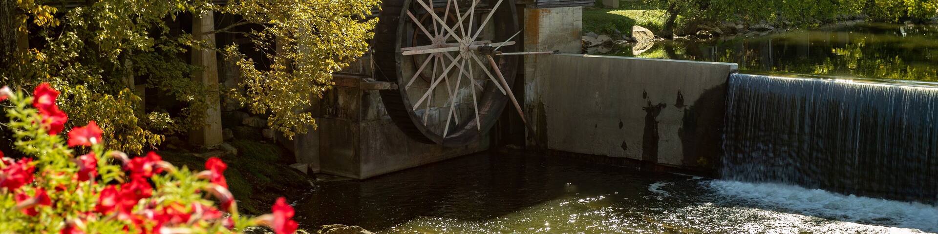 A wooden mill with a large water wheel, surrounded by lush greenery and a small waterfall.