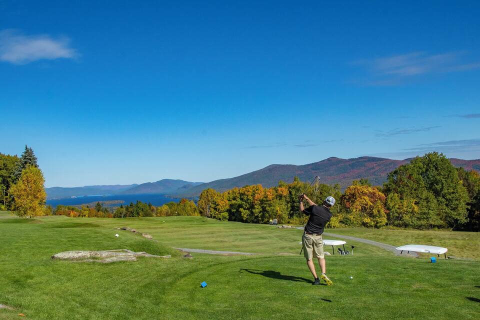 Un campo da golf con cielo azzurro, alberi autunnali e montagne sullo sfondo.