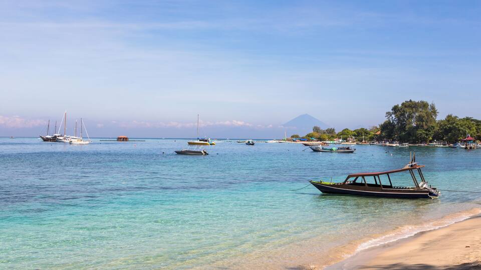 A coastal scene with boats anchored near the shore, a sandy beach, and a mountain in the background.