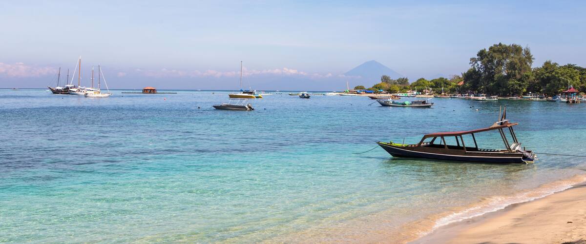A coastal scene with boats anchored near the shore, a sandy beach, and a mountain in the background.