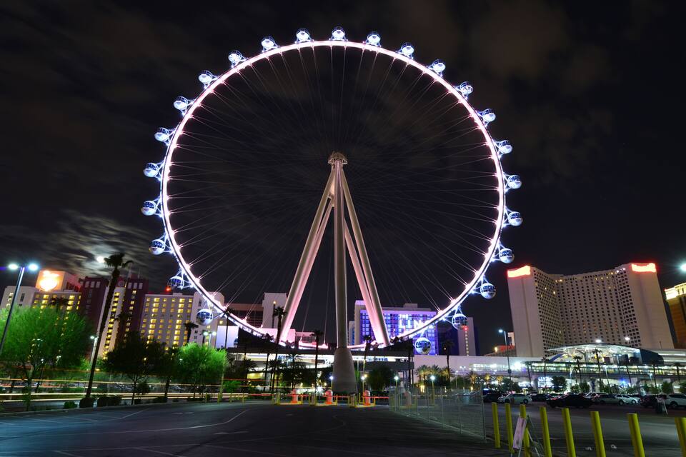 Une grande grande roue illuminée la nuit, avec des bâtiments environnants et un parking.