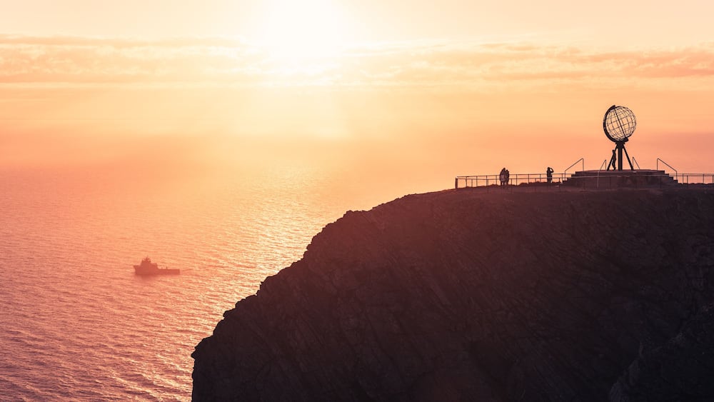 Un coucher de soleil sur une falaise côtière, avec une grande sculpture en forme de globe et un navire sur l’eau.
