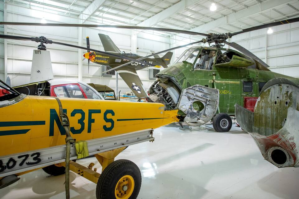 Un aereo giallo con il numero 8073 e le lettere “N3FS” sul lato, un elicottero verde e un aereo militare d’epoca in un hangar.
