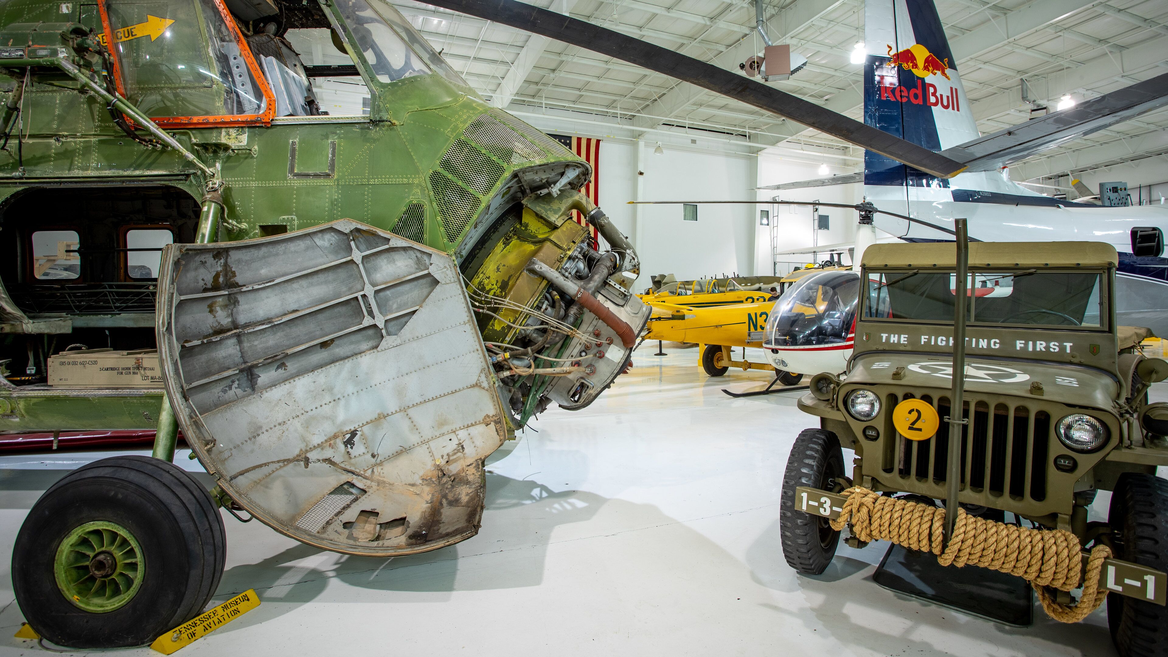 A military aircraft with a damaged wing, a jeep labeled "The Fighting First," and a Red Bull logo on another aircraft.