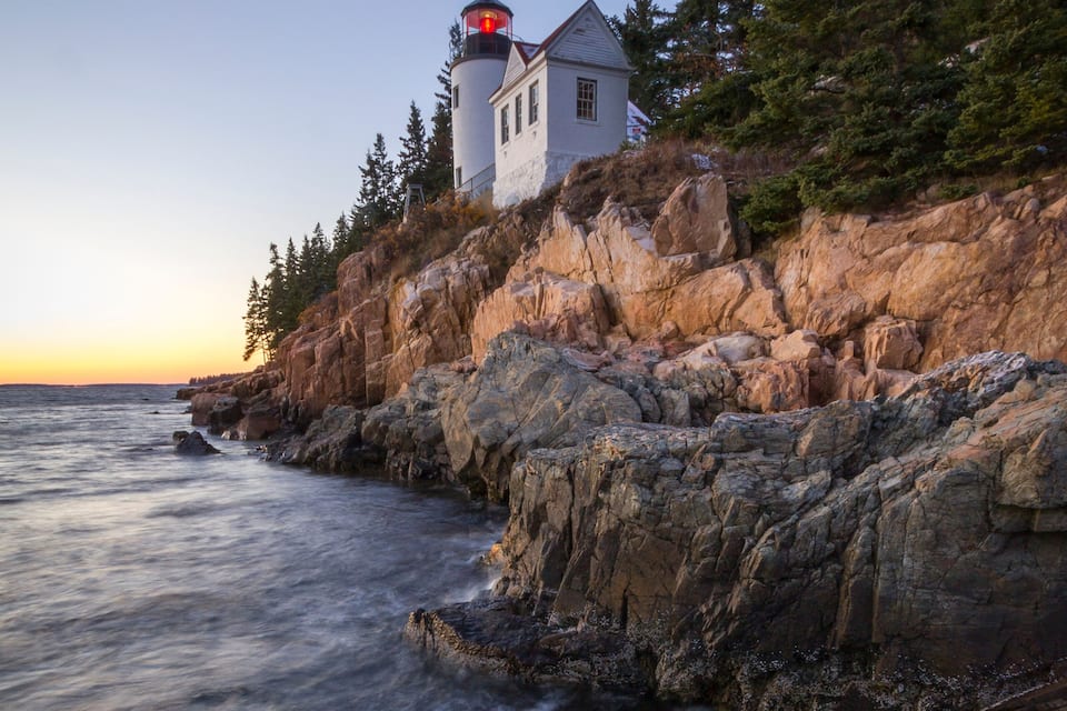 A lighthouse on a rocky cliff by the sea, with trees and a clear sky.