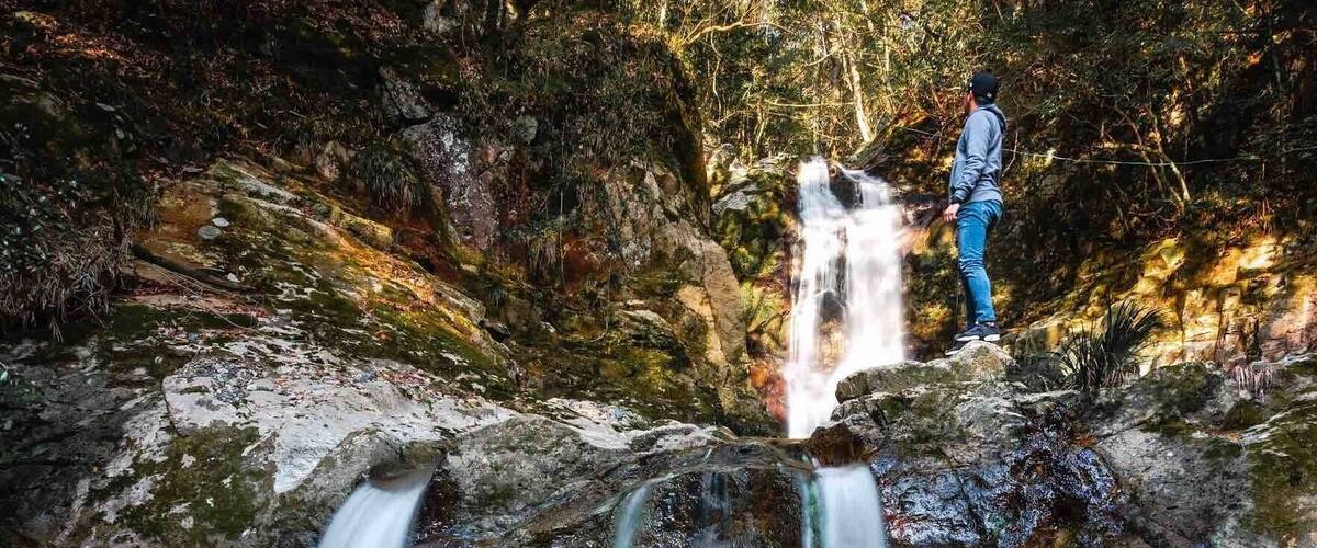 A waterfall cascading over rocks in a forested area with a person standing on a rock above the water.