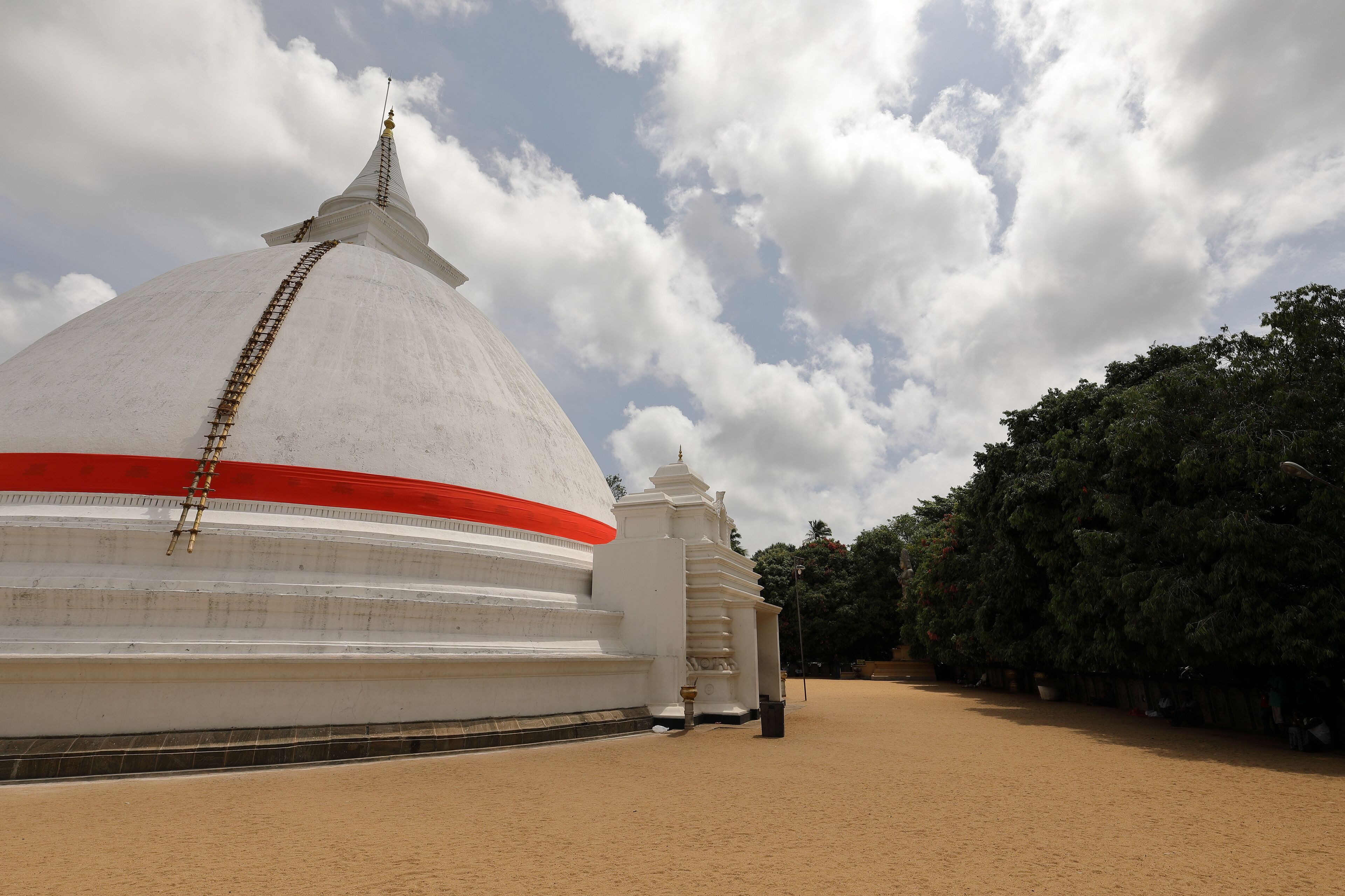 A large white stupa with a red stripe, a smaller stupa, and a tree-lined path.