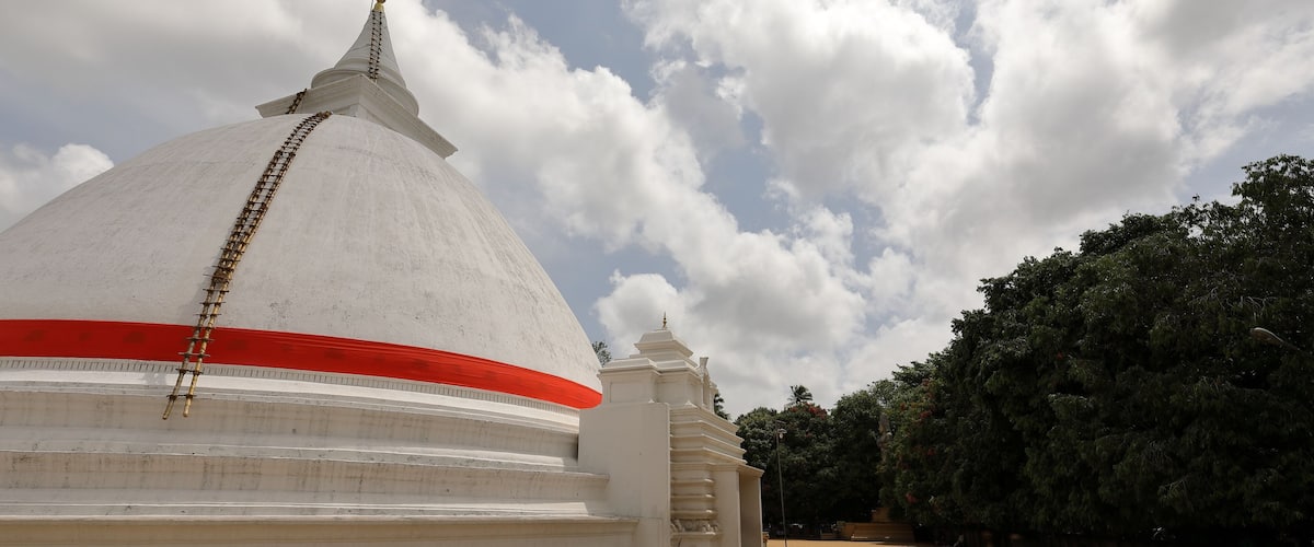 A large white stupa with a red stripe, a smaller stupa, and a tree-lined path.