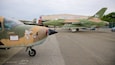 Des avions militaires sont stationnés sur un aérodrome, et on aperçoit en arrière-plan un bâtiment portant l’inscription « Musée de l’aviation américaine ».