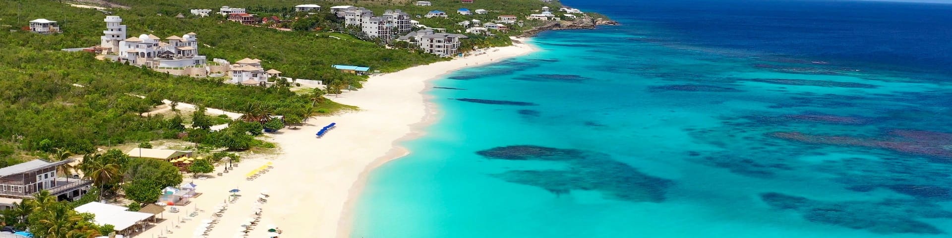A coastal resort with a white sandy beach, turquoise waters, and a backdrop of lush greenery and buildings.