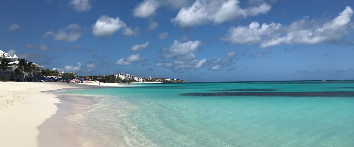 A beach with clear turquoise waters, white sand, and buildings in the background.