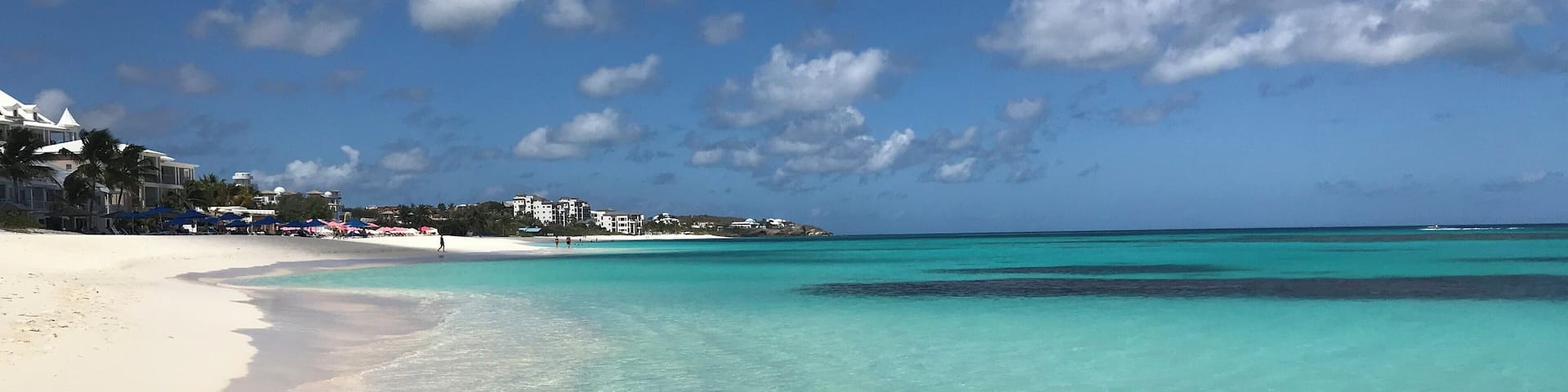 A beach with clear turquoise waters, white sand, and buildings in the background.