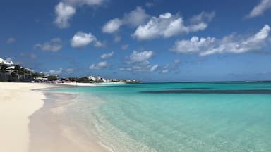 Una playa con aguas turquesas cristalinas, arena blanca y edificios al fondo.