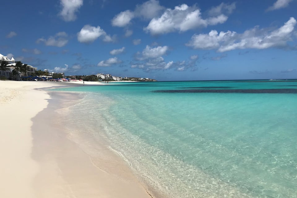 A beach with clear turquoise waters, white sand, and buildings in the background.