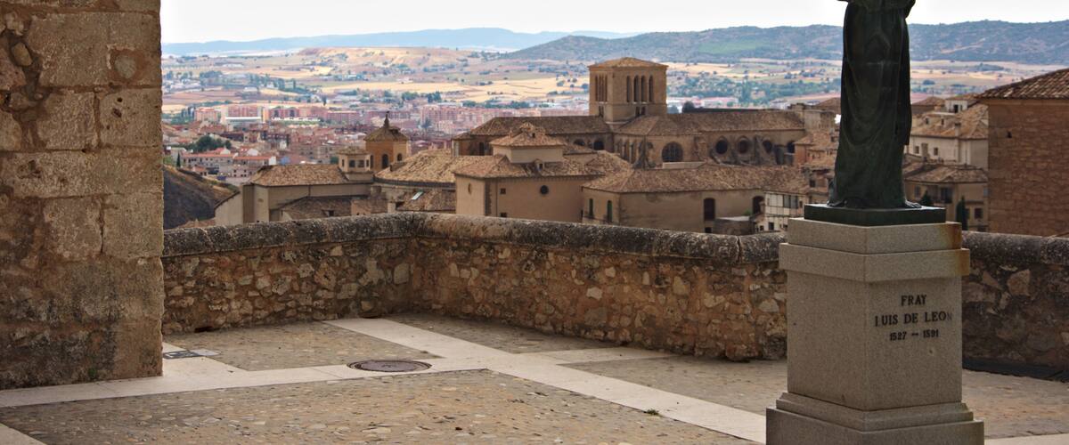 A statue of a historical figure in a square with stone walls and a view of a town in the background.