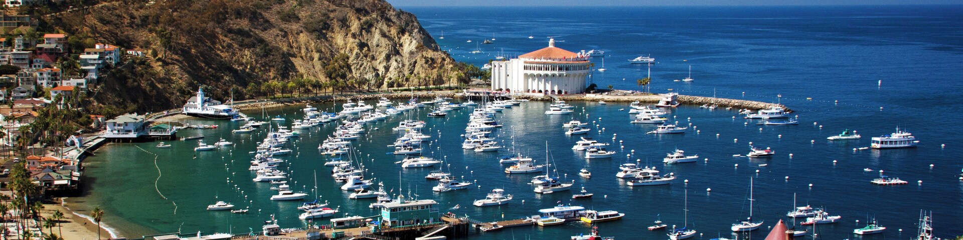 A coastal town with a marina filled with boats, a lighthouse, and a hill with buildings.