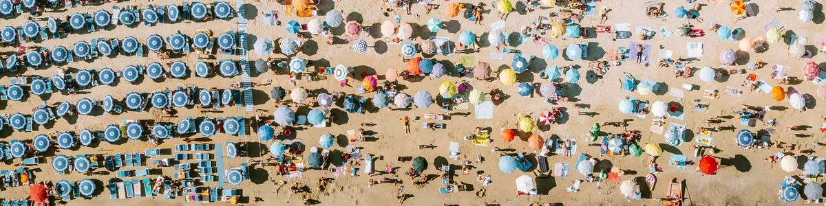 An aerial view of a beach with numerous umbrellas and sun loungers, a clear blue sea, and a road with vehicles and buildings in the background.