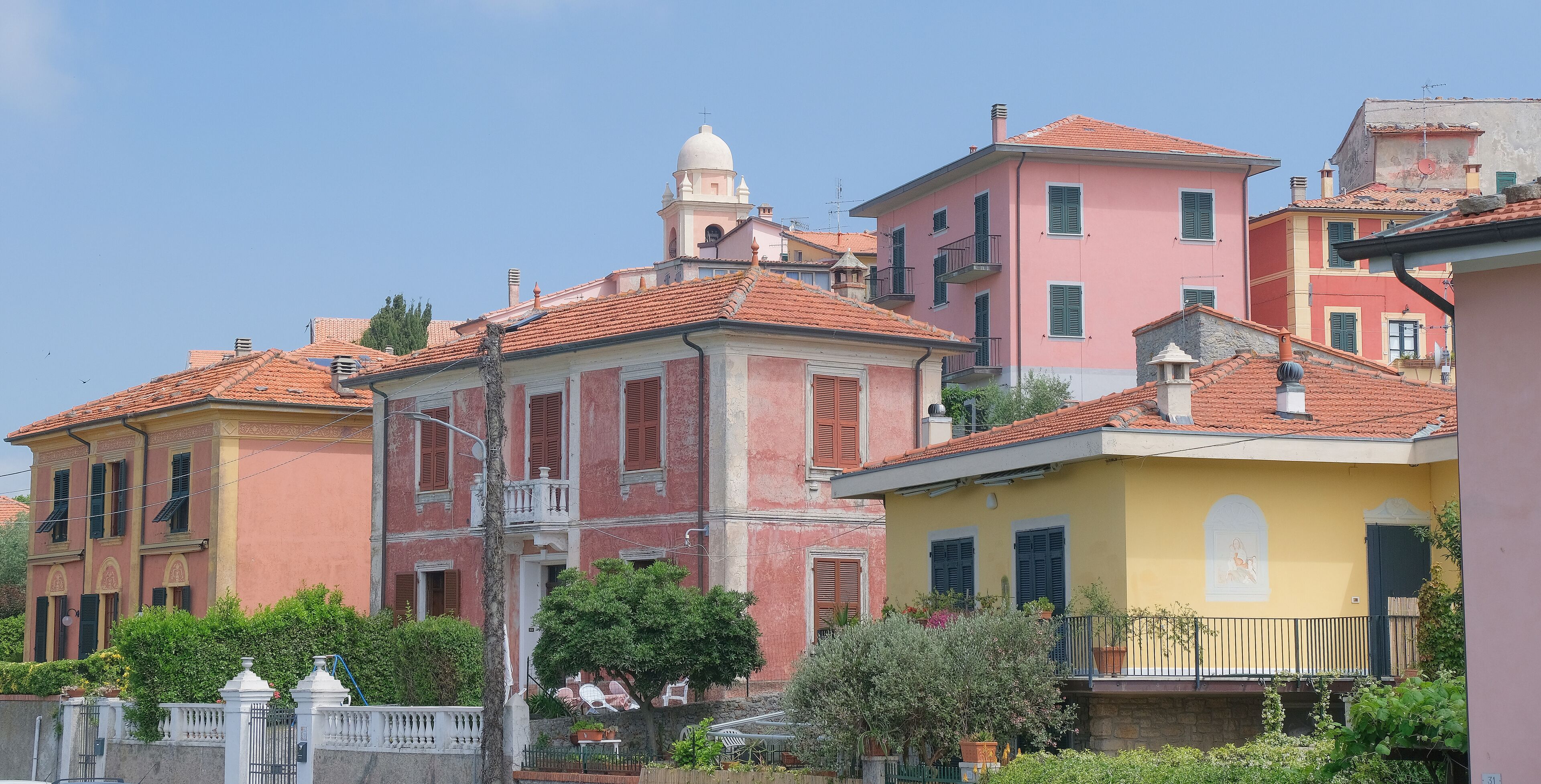 A row of colorful buildings with red-tiled roofs and balconies, surrounded by greenery.