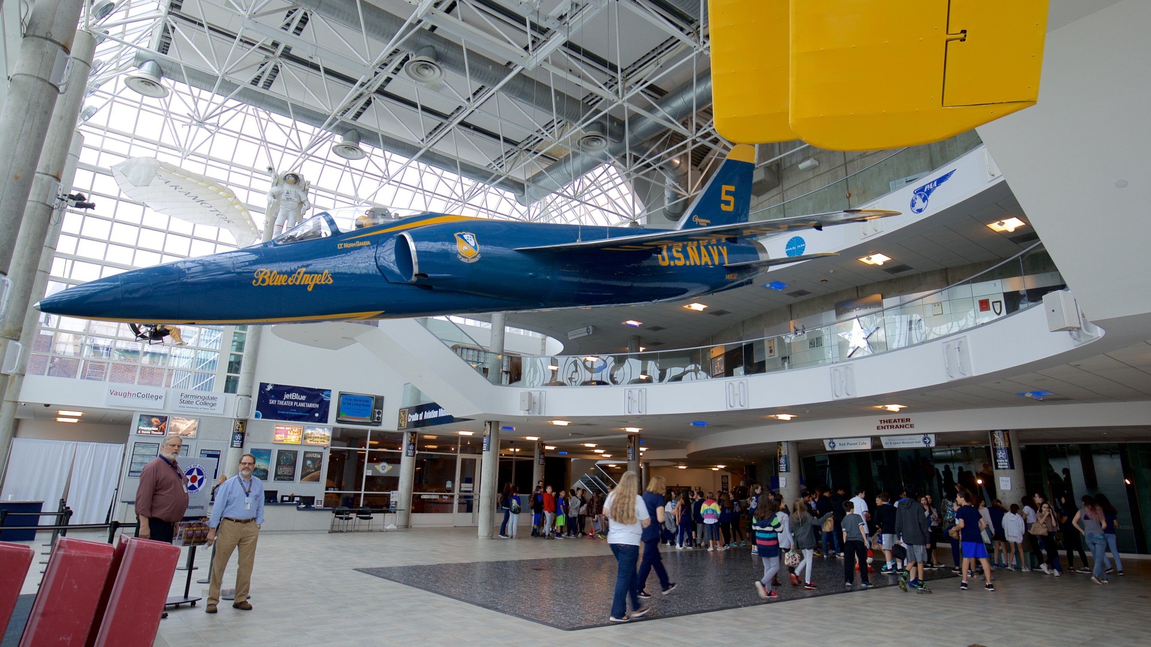 A blue and yellow airplane with 'US NAVY' and 'Blue Angels' written on it, suspended in a building with a glass ceiling.