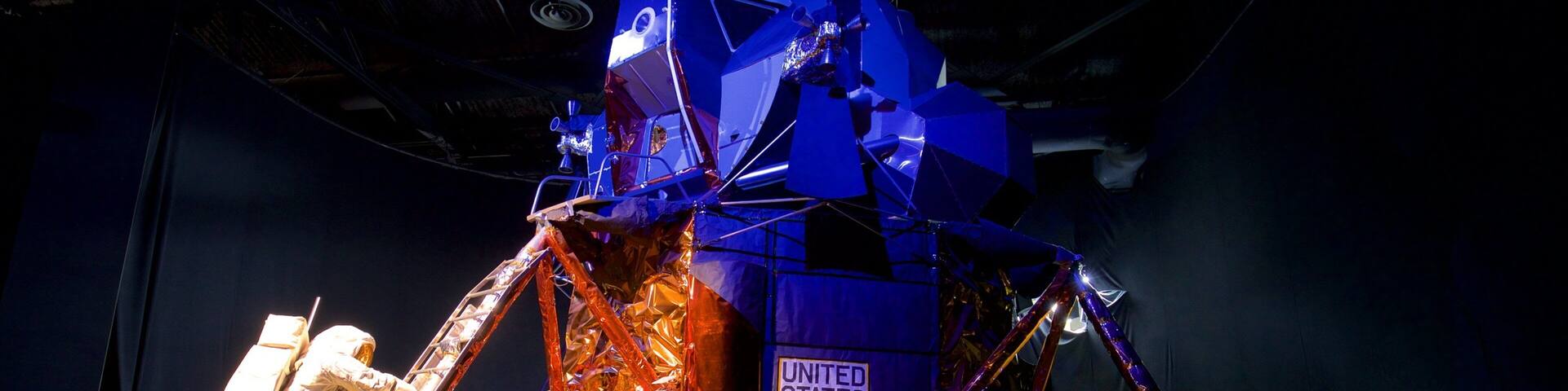 A large, metallic space shuttle model with the United States flag, surrounded by a sandy area and a person in a spacesuit.
