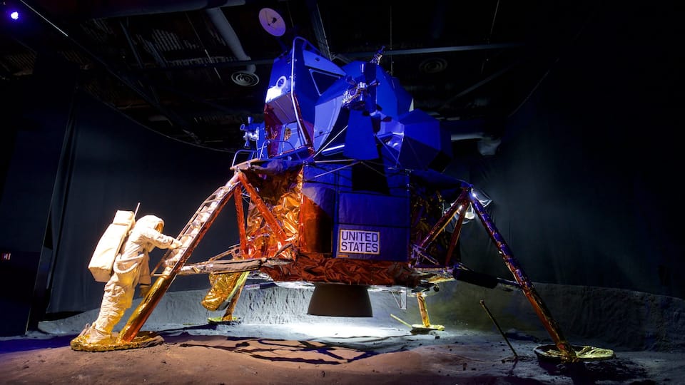 A large, metallic space shuttle model with the United States flag, surrounded by a sandy area and a person in a spacesuit.