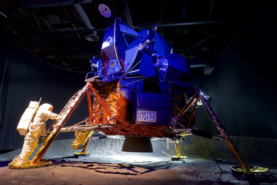 A large, metallic space shuttle model with the United States flag, surrounded by a sandy area and a person in a spacesuit.