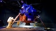 Ein großes, metallisches Space-Shuttle-Modell mit der Flagge der Vereinigten Staaten, umgeben von sandigem Gelände und einer Person im Raumanzug.