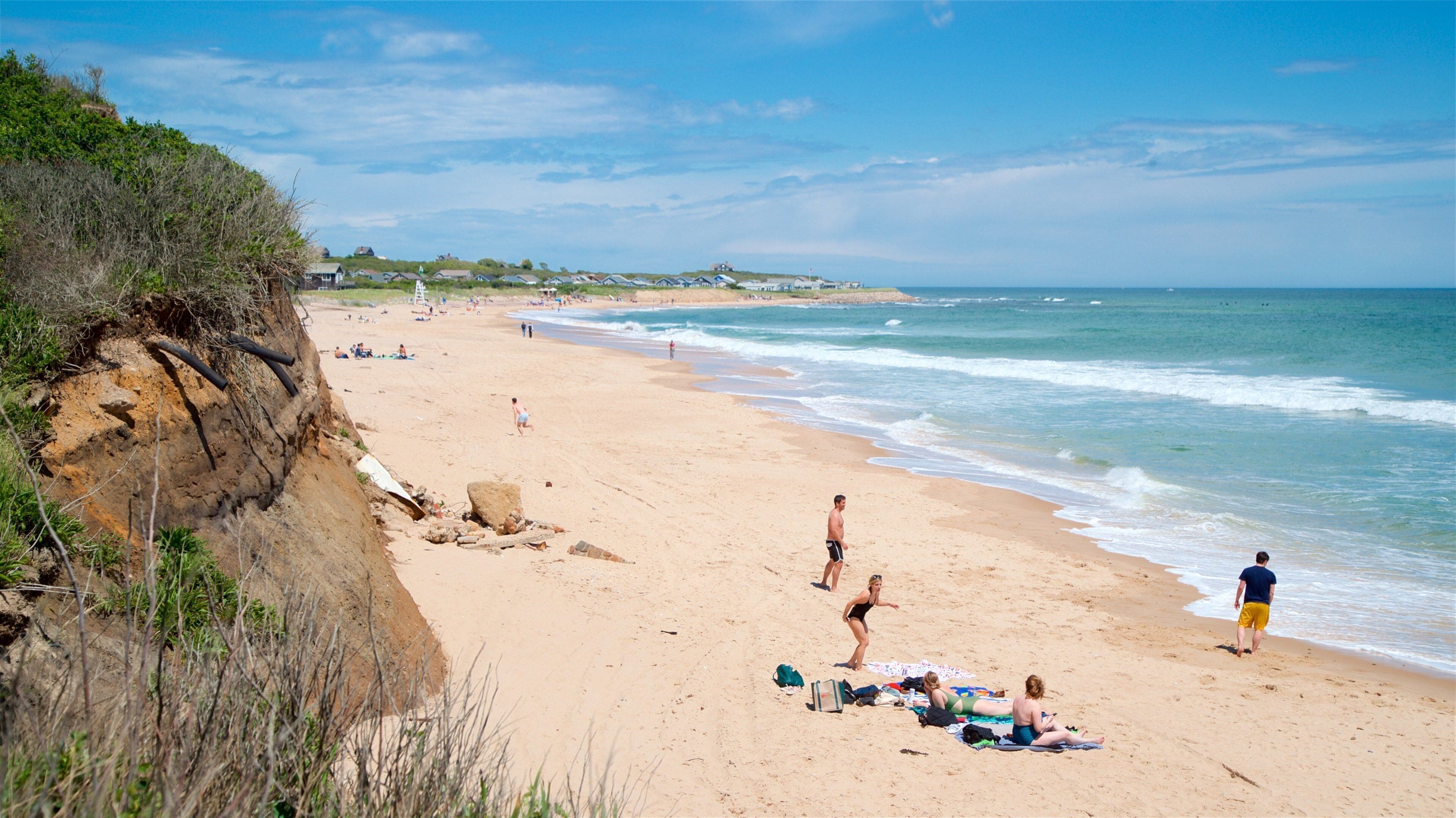 A sandy beach with people sunbathing and swimming, a cliff with vegetation, and a calm sea.