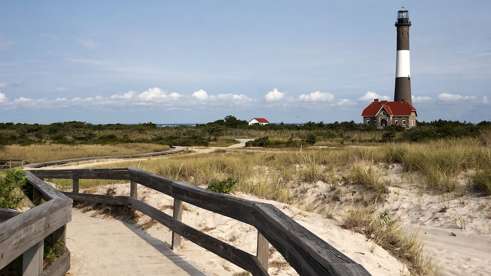 A lighthouse, a wooden boardwalk, and a red-roofed building in a coastal landscape.