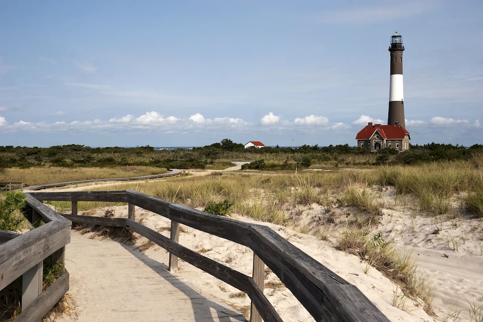 A lighthouse, a wooden boardwalk, and a red-roofed building in a coastal landscape.