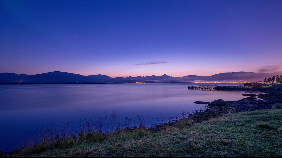 A calm lake at dusk with mountains in the background and a pier extending into the water.