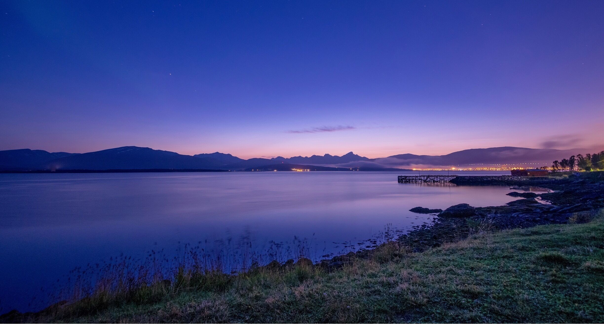 A calm lake at dusk with mountains in the background and a pier extending into the water.