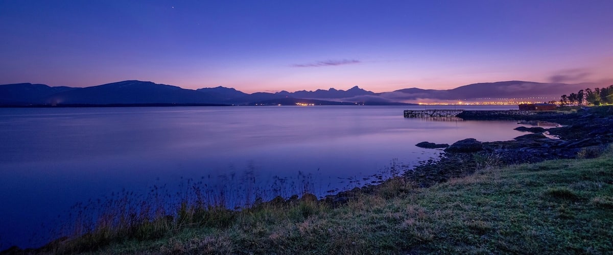 A calm lake at dusk with mountains in the background and a pier extending into the water.