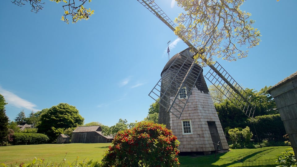 A traditional windmill with a wooden structure and a lattice roof, surrounded by lush greenery and a clear blue sky.