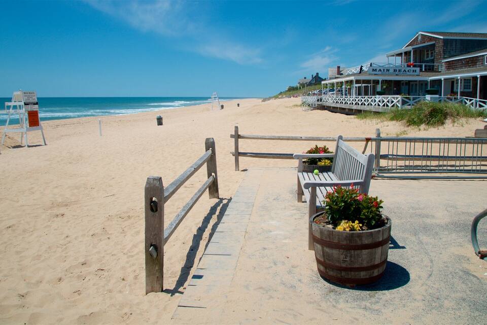 Ein Strand mit einer Holzbank, einem Rettungsschwimmerstand und einem Gebäude mit der Aufschrift 'Main Beach'.