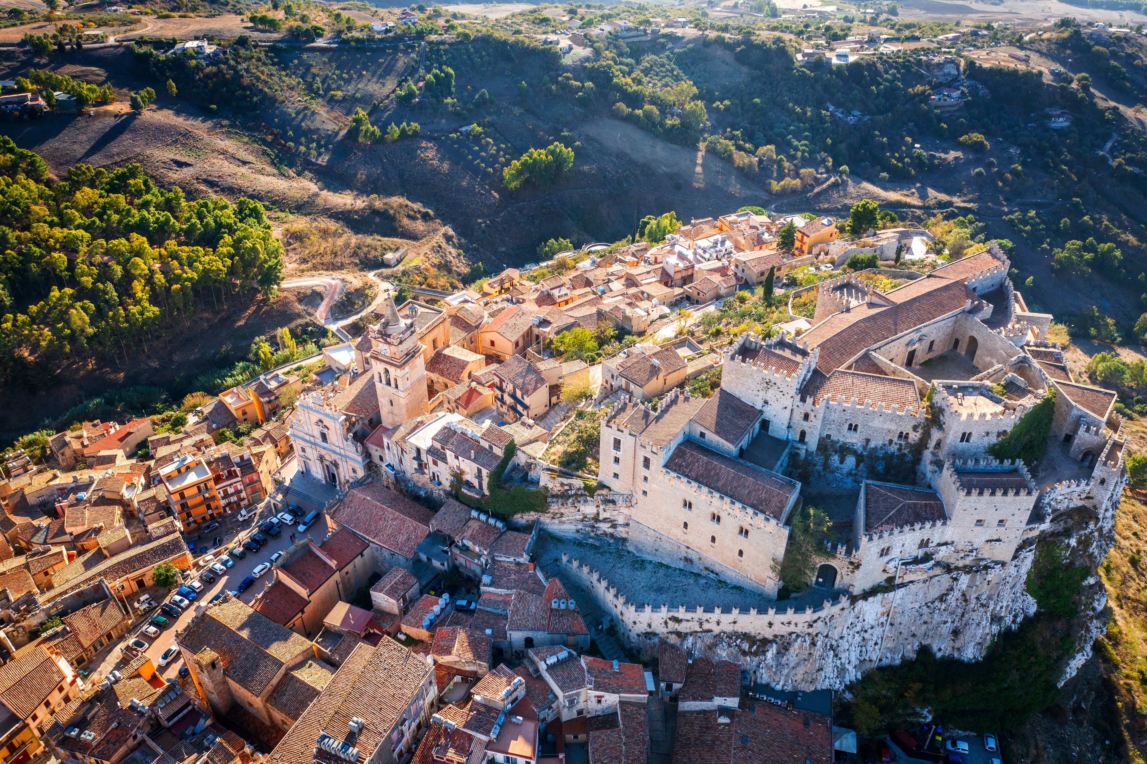 An aerial view of a historic town with a prominent castle on a hill, surrounded by terraced vineyards and a valley.