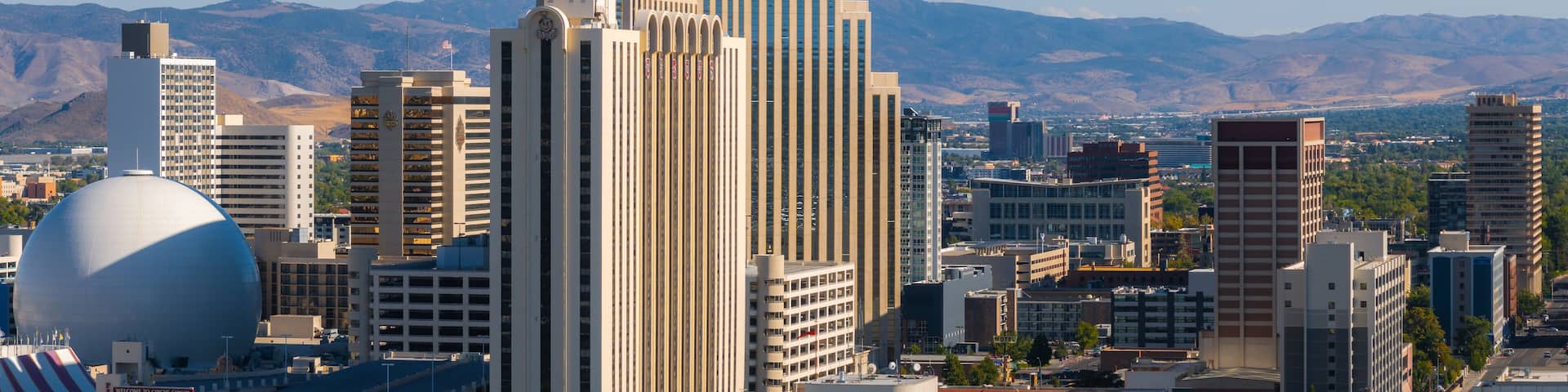 Reno, Nevada's skyline is seen from above, featuring the silver dome of the National Bowling Stadium and the Sierra Nevada mountains in the background.