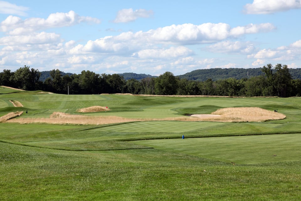 Um campo de golfe com colinas verdes ondulantes, áreas de areia e uma encosta arborizada ao fundo.