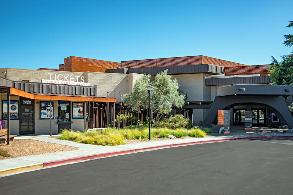 A building with a 'Tickets' sign, a box office, and a landscaped area with trees and shrubs.