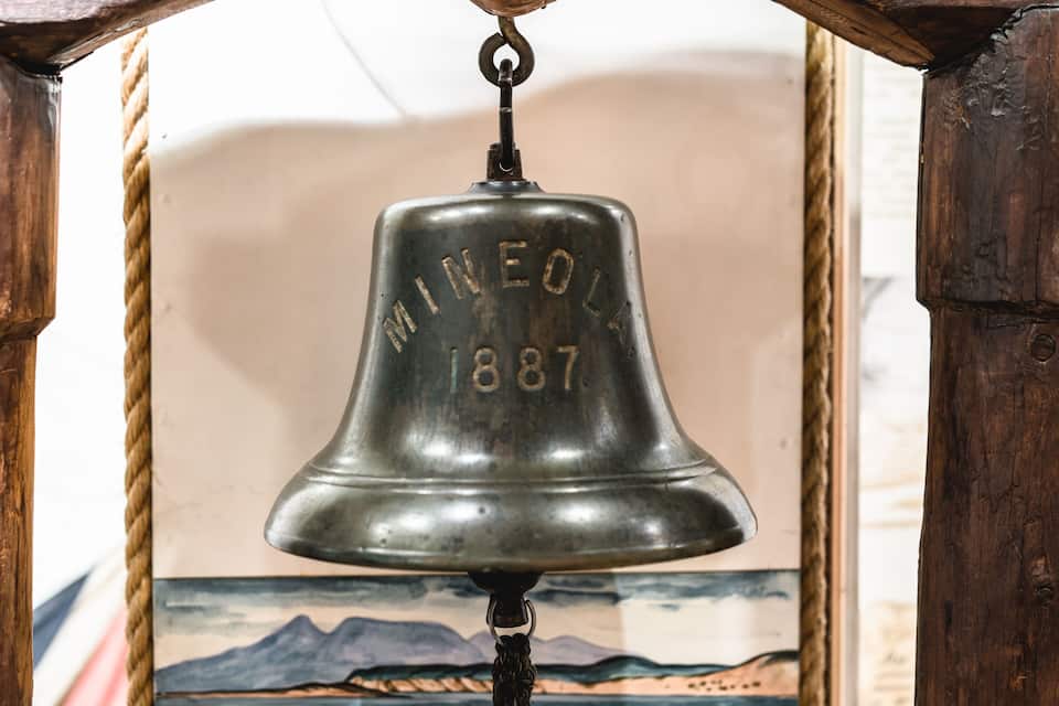 A vintage bell with the inscription "MINEOLA 1887" hanging from a wooden structure.