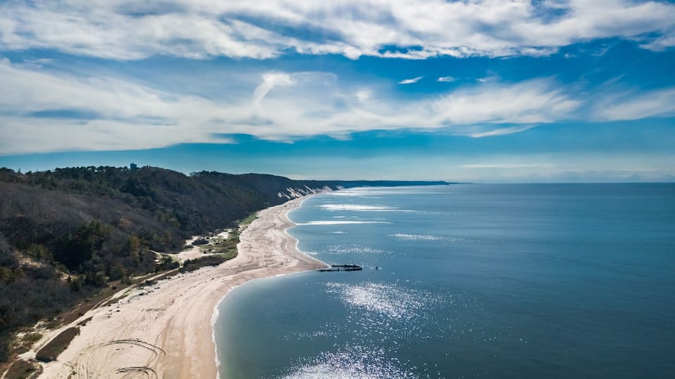 A coastal landscape with a sandy beach, a pier extending into the water, and a forested hillside.