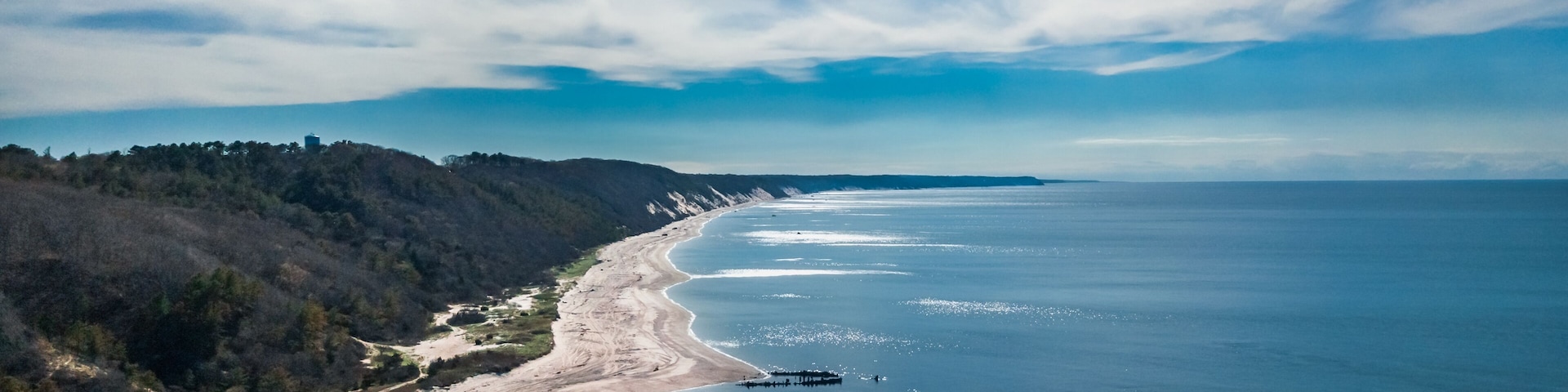 A coastal landscape with a sandy beach, a pier extending into the water, and a forested hillside.